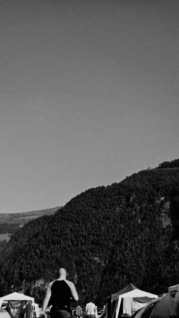 Silhouetted person with a shaved head in a sleeveless shirt walks toward a forested mountain and tents at a campsite (black and white).